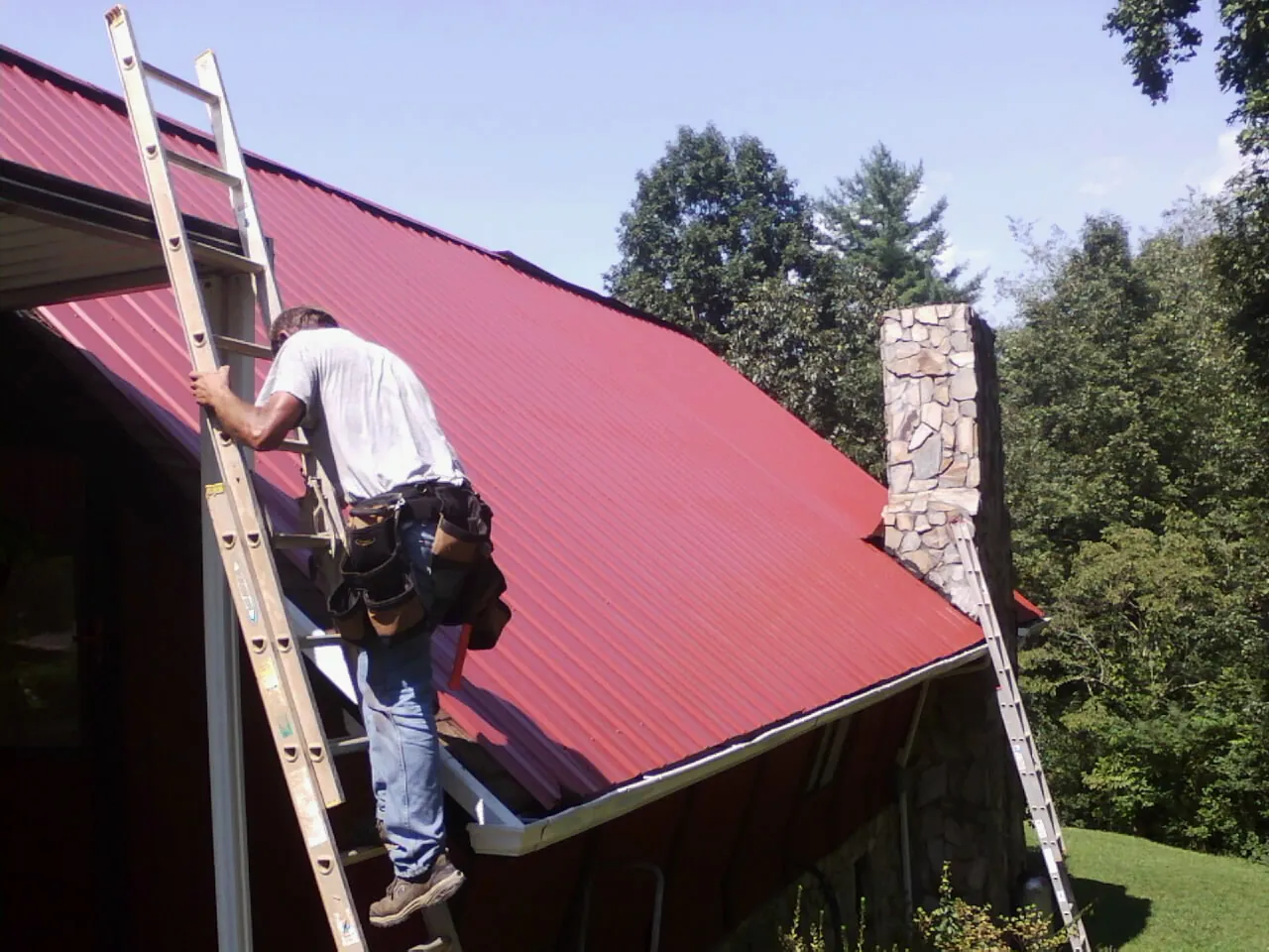 Roofer on a ladder beside a red metal roof and stone chimney — Soffit & Fascia Repair by Roofing Brothers in Vantage